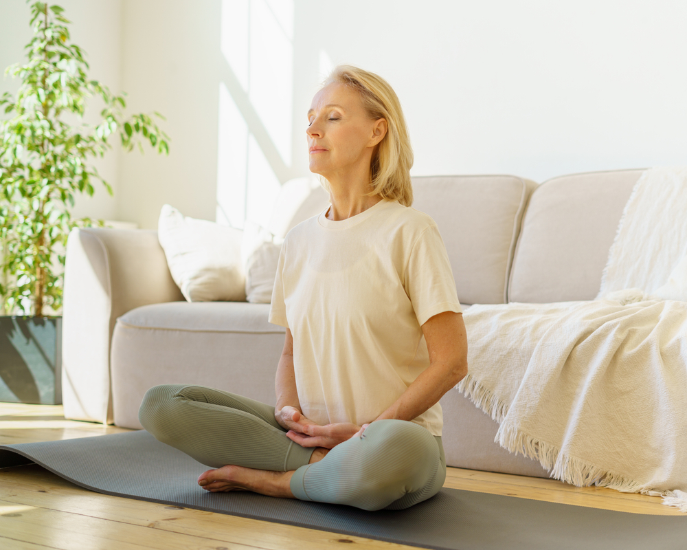 Peaceful senior woman in lotus position meditation with closed eyes at home while sitting on yoga mat on floor, full length. Calm elderly lady practicing meditation techniques and yoga indoors